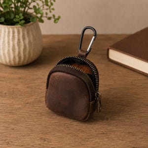 May include: A small, brown leather pouch with a zip closure and a black carabiner clip. The pouch is open, revealing a brown interior. The pouch is on a wooden surface.