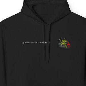 Puede incluir: Sudadera con capucha negra con bolsillo delantero. La sudadera presenta el texto "sudo restart universe" y un gráfico de un bicho verde con gafas y sombrero, trabajando en un portátil.