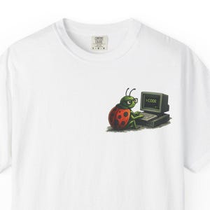 Könnte beinhalten: Weißes T-Shirt mit einem Cartoon-Marienkäfer und Computer-Grafik. Der Marienkäfer ist rot mit schwarzen Punkten, und der Computerbildschirm zeigt das Wort "CODE". Das Design befindet sich im linken Brustbereich.