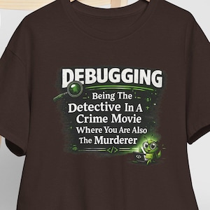 May include: A brown t-shirt featuring the text "DEBUGGING" and the phrase "Being The Detective In A Crime Movie Where You Are Also The Murderer" in white and green. A white baseball cap rests on a wooden chair.