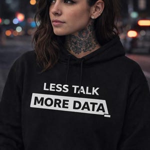 Puede incluir: Sudadera con capucha negra con el texto blanco "LESS TALK MORE DATA". La persona lleva la sudadera con capucha y tiene un portátil. El fondo es una escena urbana borrosa con luces.