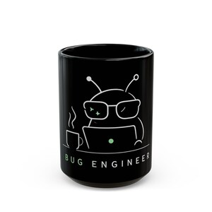 May include: Black ceramic mug with a white cartoon bug wearing glasses and working on a laptop. The mug has the words "BUG ENGINEER" in green and white. A small cup of coffee is also depicted.