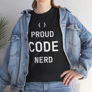 以下が含まれることがあります： 白文字で「{ } PROUD CODE NERD」と書かれた黒いTシャツ。青いデニムジャケットとブルージーンズを着用しています。Tシャツのデザインは、単語の上に波括弧が特徴です。