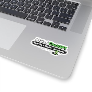 May include: A white sticker with the text "I Asked ChatGPT Now I'm A Senior Developer" in black and green. The sticker also features a cartoon frog and code symbols. The sticker is placed on a silver laptop.