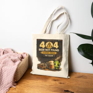 Puede incluir: Un bolso tote beige con un diseño humorístico. El diseño presenta un mensaje de error de computadora: "404 BEER NOT FOUND SYSTEM CRASH TRY AGAIN". Debajo del texto hay ilustraciones de jarras de cerveza y un triste personaje de dibujos animados verde.