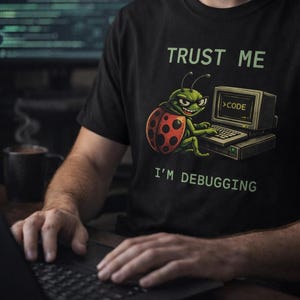 May include: Black t-shirt featuring the text "TRUST ME I'M DEBUGGING" and a cartoon ladybug character. The ladybug is sat in front of a computer. The person in the image is using a laptop.