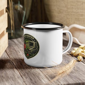 Pode incluir: Caneca esmaltada branca com borda e alça pretas. A caneca apresenta um gráfico circular com o texto "BUG HUNTER" e uma ilustração de um inseto verde com um computador. A caneca está sobre uma superfície de madeira.