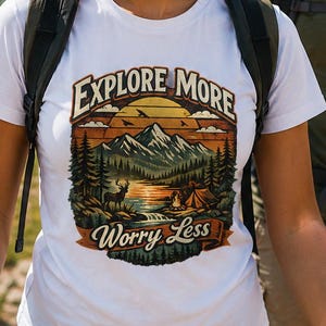 Könnte beinhalten: Ein weißes kurzärmeliges T-Shirt mit einem Grafikdruck einer Berglandschaft mit einem Hirsch, einem Zelt und einem Lagerfeuer. Der Text "EXPLORE MORE" ist darüber gewölbt und "Worry Less" steht auf einem Banner am unteren Rand.