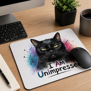 Könnte beinhalten: Weißes Mauspad mit einer Illustration einer schwarzen Katze mit gelben Augen und dem Text "I AM Unimpressed". Die Katze ist von bunten Farbspritzern umgeben. Eine schwarze Computermaus befindet sich rechts.