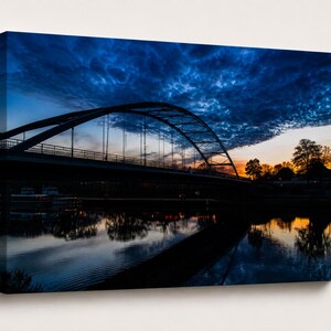 Könnte beinhalten: Leinwanddruck einer Brücke über Wasser in der Abenddämmerung. Die Brücke hat ein Bogendesign und ist vor einem lebendigen Himmel mit Blau-, Orange- und Gelbtönen silhouettiert. Das Wasser spiegelt die Brücke und den Himmel wider.