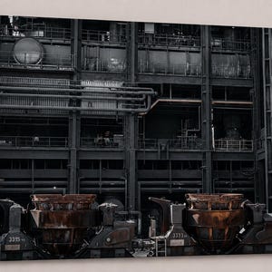 Könnte beinhalten: Schwarz-Weiß-Industriefotografie eines Fabrikinneren. Das Bild zeigt große, verrostete Metallmaschinen im Vordergrund, mit Rohren und Strukturelementen. Die Gesamtästhetik ist dunkel und körnig.
