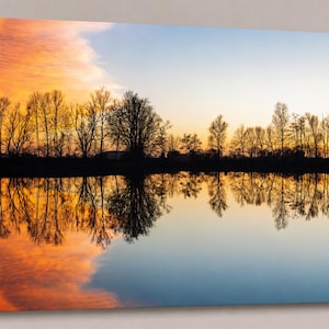 Könnte beinhalten: Ein Leinwanddruck einer ruhigen Landschaft mit einem See, der einen lebendigen Sonnenuntergang widerspiegelt. Der Himmel wechselt von Orange zu Blau, mit silhouettierten Bäumen am Horizont. Das Wasser spiegelt Himmel und Bäume wider und erzeugt ein symmetrisches Bild.