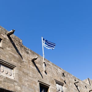 Könnte beinhalten: Ein Steinbau mit einer griechischen Flagge, die im Wind vor einem klaren blauen Himmel weht. Das Gebäude hat mehrere Fenster und dekorative Elemente, mit Schatten.