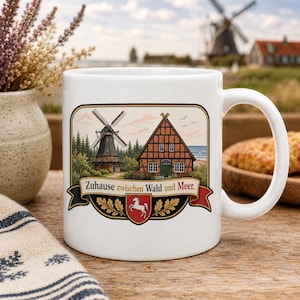 Può includere: Una tazza in ceramica bianca con un'illustrazione colorata di un mulino a vento e una casa a graticcio vicino al mare. Sotto la scena, un banner mostra il testo "Zuhause zwischen Wald und Meer" sopra uno scudo con l'emblema di un cavallo bianco.
