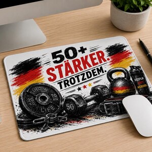 Könnte beinhalten: Weißes Mauspad mit Fitness-Motiv, mit Gewichten, einer Kettlebell und dem Text "50+ STÄRKER. TROTZDEM." in Schwarz und Rot. Das Design integriert die Farben der deutschen Flagge. Eine weiße Maus und andere Büromaterialien.