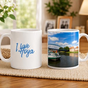 Puede incluir: Dos tazas de cerámica blanca. Una taza tiene el texto "I love Hoya" en escritura azul. La otra taza presenta una imagen escénica de un barco en un río bajo un cielo azul con un puente y edificios al fondo.