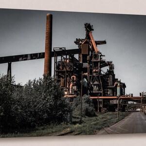 Könnte beinhalten: Leinwanddruck einer Industrielandschaft mit einer großen Fabrik und einem hohen Backsteinkamin. Die Szene wird von Grau-, Braun- und Orangetönen dominiert, mit einer Straße, die in die Ferne führt.