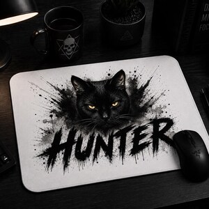 Könnte beinhalten: Rechteckiges weißes Mauspad mit einem schwarzen Katzendesign und dem Wort "HUNTER" in Schwarz. Die Katze hat gelbe Augen und ist von schwarzer Farbspritzern umgeben. Eine schwarze Computermaus befindet sich neben dem Mauspad.