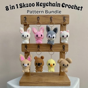 Op de afbeelding: Een houten displaystandaard met acht gehaakte dieren sleutelhangers. De sleutelhangers zijn een wit konijn, roze konijn, grijze wolf, witte kat, bruine vos, bruine beer, gele kuiken en bruine hond. De tekst luidt "8 in 1 Skzoo Keychain Crochet Pattern Bundle".
