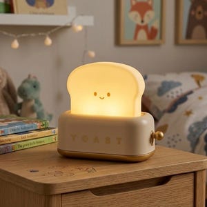 May include: A whimsical, cream-colored night light shaped like a toaster with a smiling piece of toast. The word "TOAST" is printed on the front. The night light has a gold-colored base and a small knob on the side.