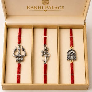 May include: A boxed set of three Rakhi bracelets. Each bracelet features a red cord and a silver-toned charm. The charms depict a trident, the Om symbol, and a square pendant with text. The box is labelled "RAKHI PALACE".