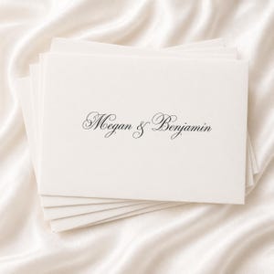 Op de afbeelding: Een stapel ivoorkleurig briefpapier met de namen "Megan & Benjamin" in elegant zwart schrift. Het briefpapier is gerangschikt op een witte satijnen ondergrond, wat een zachte, luxueuze uitstraling creëert.
