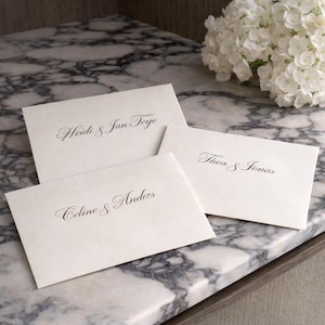 Puede incluir: Una taza de cerámica blanca con asa negra y el texto "But First, Coffee" en negrita negra. La taza se encuentra sobre un fondo borroso, sugiriendo un ambiente acogedor.