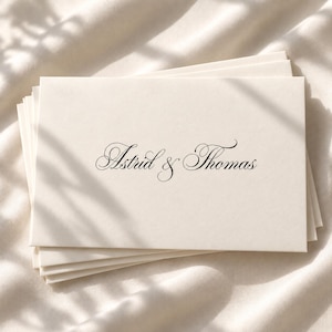 May include: A stack of cream-coloured rectangular envelopes featuring the names Astrid & Thomas printed in a black, elegant, flowing script font centred on the front of the top envelope. The paper has a smooth, matte finish and clean edges.