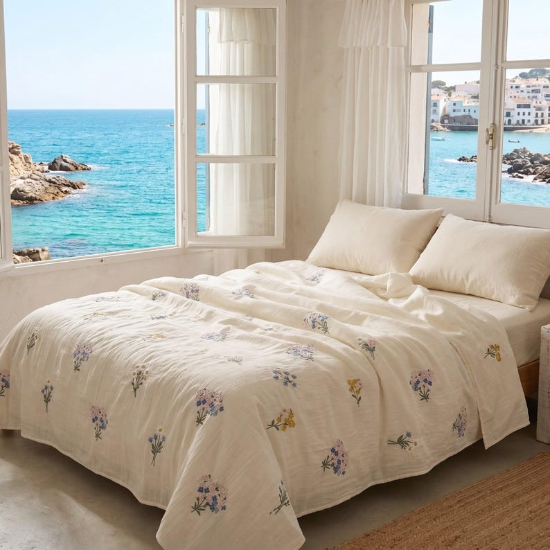 May include: A cream-colored bedspread and two matching pillowcases featuring a scattered pattern of small, embroidered floral bouquets in shades of blue, yellow, and purple. The bedding is draped over a bed frame positioned near a large window.