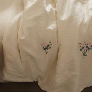 May include: A cream-colored fabric sheet featuring delicate embroidered floral motifs. The textile has a subtle, crinkled texture and displays two distinct small bouquets of flowers in shades of pink, white, green, and blue stitched onto the surface.