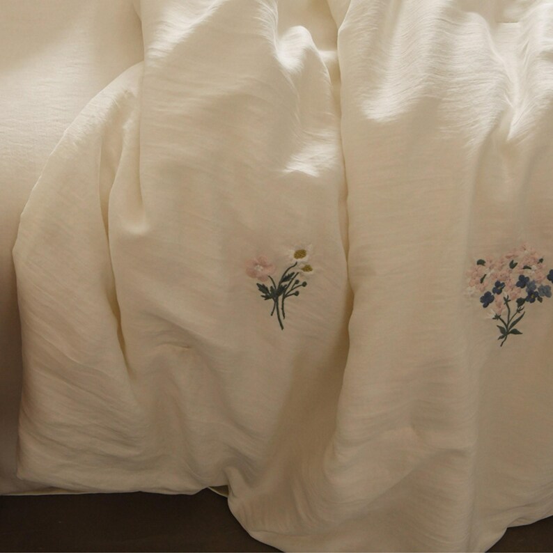 May include: A cream-colored fabric sheet featuring delicate embroidered floral motifs. The textile has a subtle, crinkled texture and displays two distinct small bouquets of flowers in shades of pink, white, green, and blue stitched onto the surface.