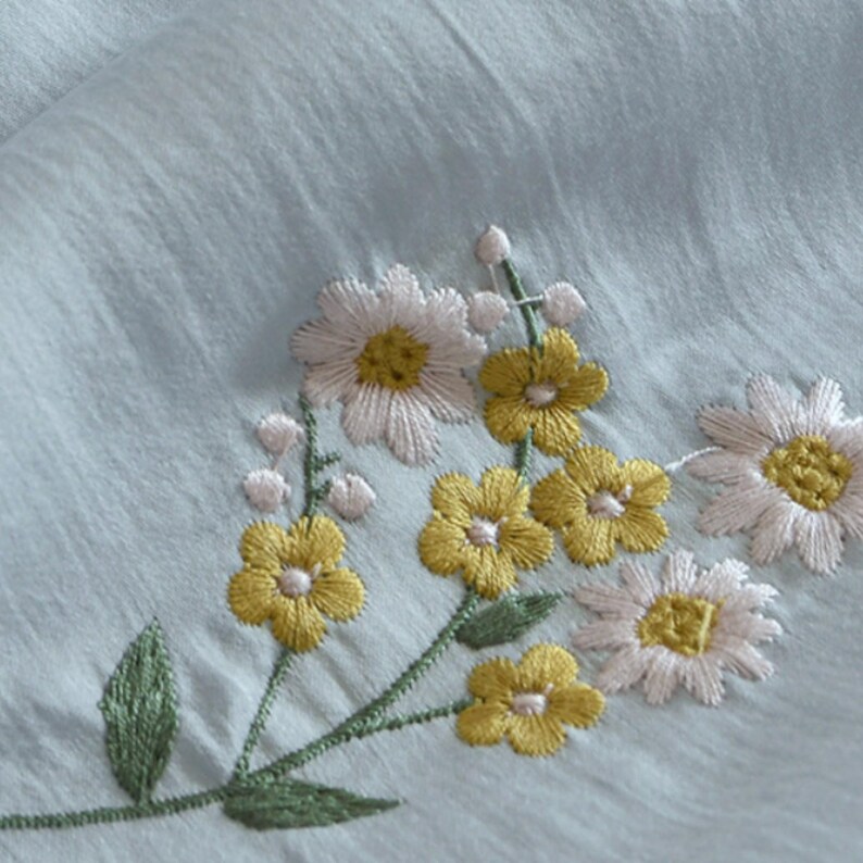 May include: A light blue fabric featuring an embroidered floral design. The embroidery consists of white daisies with yellow centers, small yellow five-petaled flowers, and green stems with leaves. The fabric has a subtle, crinkled, and textured surface.