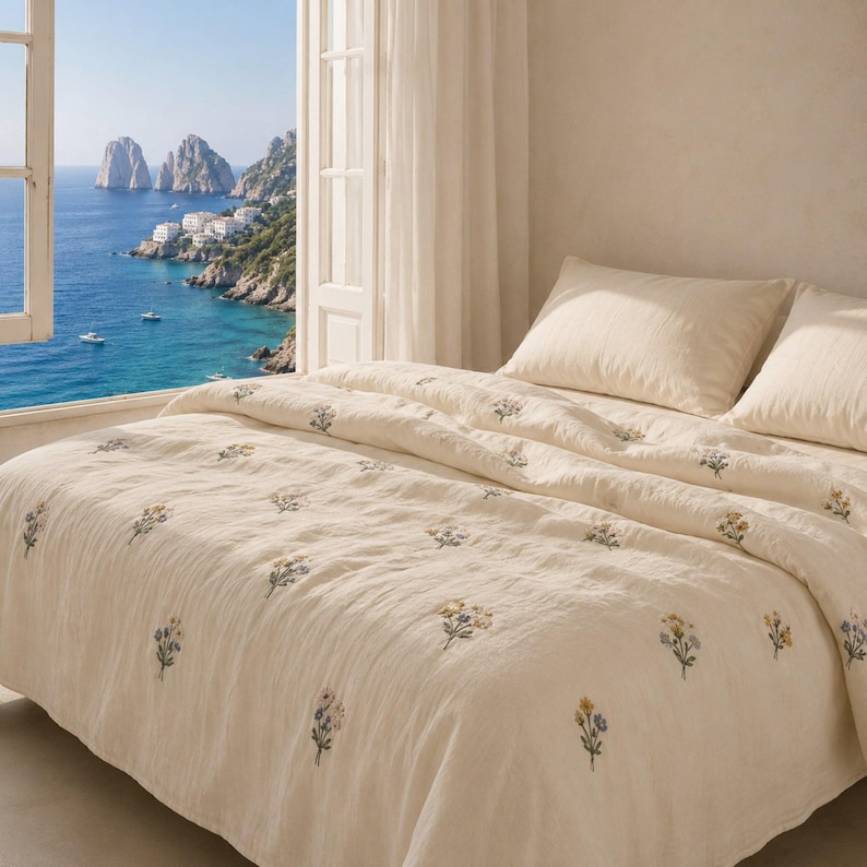 May include: A cream-colored bedspread and two matching pillowcases featuring a scattered pattern of small, embroidered floral bouquets in shades of blue, yellow, and green. The bedding has a textured fabric appearance and is neatly arranged on a bed.