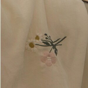 May include: A cream-colored fabric garment featuring an embroidered floral design. The embroidery consists of two white daisies with yellow centers, a single light pink flower, and thin green stems with small leaves and buds.