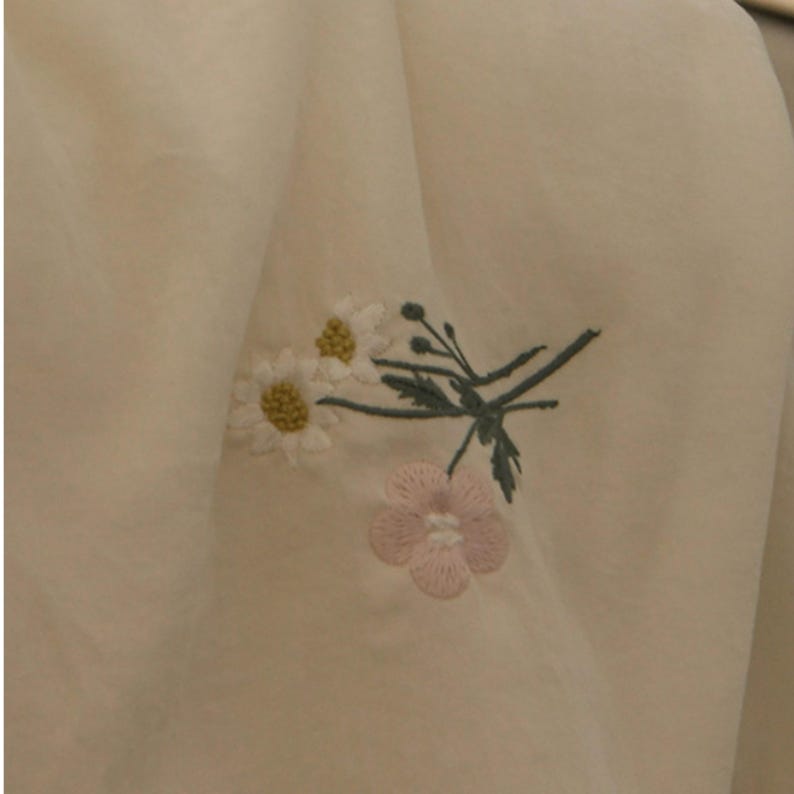 May include: A cream-colored fabric garment featuring an embroidered floral design. The embroidery consists of two white daisies with yellow centers, a single light pink flower, and thin green stems with small leaves and buds.
