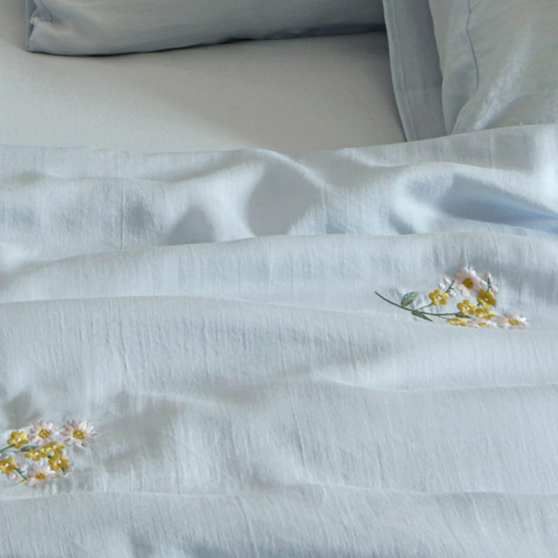 May include: A light blue bed sheet featuring small embroidered clusters of white and yellow flowers. The fabric has a visible textured weave, and the sheet is draped in soft folds, showing two distinct floral patterns on the surface.