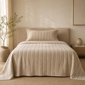 Op de afbeelding: Een beige kabelgebreid sprei en kussenset op een bed. De sprei hangt over de zijkanten van het bed. De kamer heeft een neutraal kleurenpalet, met een beige hoofdbord en een klein nachtkastje.