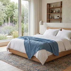 May include: A bedroom scene featuring a bed with white linens and pillows, a light blue patterned throw blanket, and a wooden bed frame. A patterned rug sits beneath the bed. A large window offers a view of a garden.