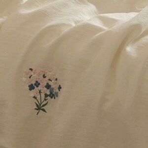 May include: A soft, off-white fabric featuring a small, delicate embroidered bouquet of flowers. The floral design includes light pink, white, and blue petals with green stems and leaves, centered on the smooth, slightly draped textile surface.