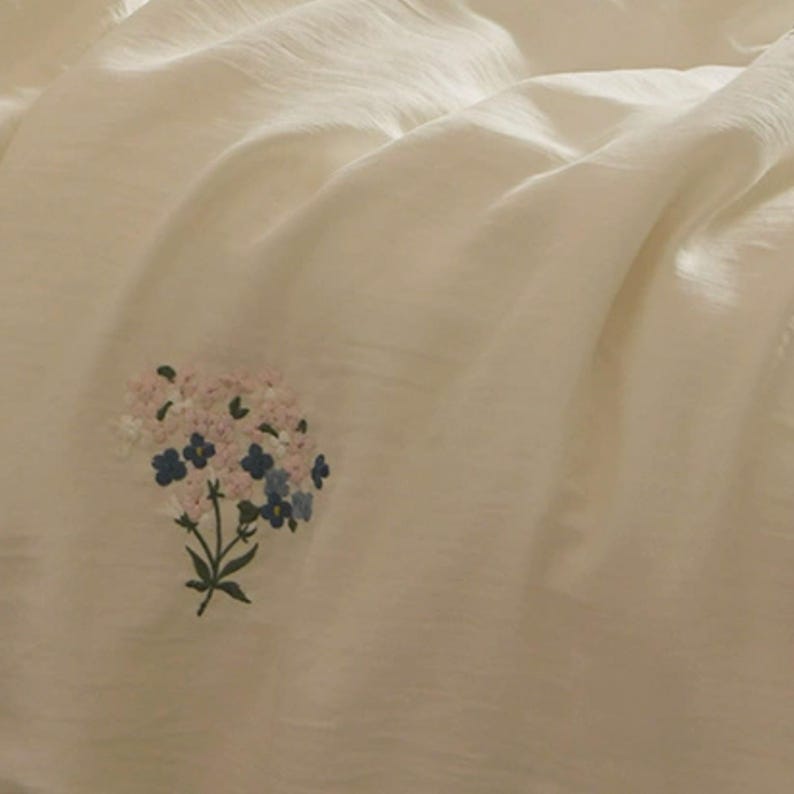 May include: A soft, off-white fabric featuring a small, delicate embroidered bouquet of flowers. The floral design includes light pink, white, and blue petals with green stems and leaves, centered on the smooth, slightly draped textile surface.