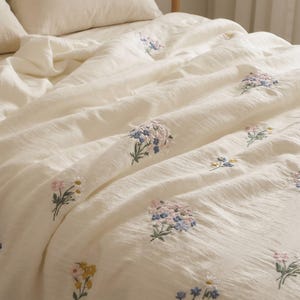 May include: A cream-colored bedspread featuring a textured fabric surface with scattered embroidered floral motifs. The embroidery includes small bouquets of flowers in shades of blue, pink, yellow, and white with green stems and leaves.