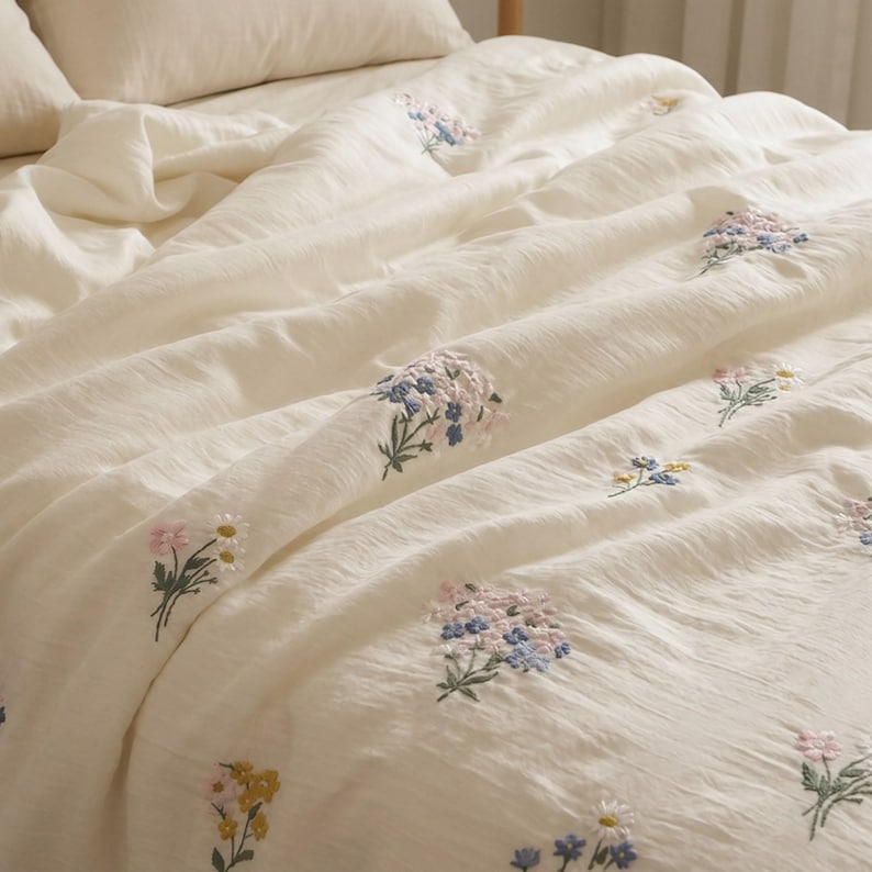 May include: A cream-colored bedspread featuring a textured fabric surface with scattered embroidered floral motifs. The embroidery includes small bouquets of flowers in shades of blue, pink, yellow, and white with green stems and leaves.