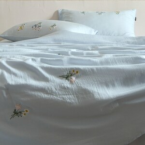 May include: A light blue bedding set consisting of a duvet cover and two pillows, all featuring a textured fabric surface and small, embroidered floral bouquets in white, yellow, and pink scattered across the light blue material.