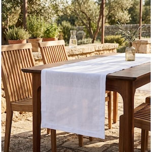 May include: A white linen table runner with a stitched border on a dark wood table. The table is set outdoors with wooden chairs, a glass carafe, and a small vase with greenery. The scene is bathed in natural light.