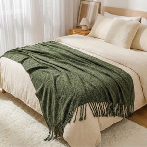 Könnte beinhalten: Eine grüne Decke mit Fransen liegt auf einem Bett. Die Decke ist einfarbig und scheint aus einem weichen Material zu bestehen. Das Bett hat cremefarbene Bettwäsche und Kissen. Eine kleine Lampe und ein gerahmtes Kunstwerk befinden sich auf dem Nachttisch.
