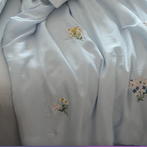 May include: A light blue fabric sheet or blanket featuring scattered embroidered floral motifs. The embroidery consists of small clusters of white, yellow, and pink flowers with green stems and leaves, distributed across the smooth, draped textile surface.