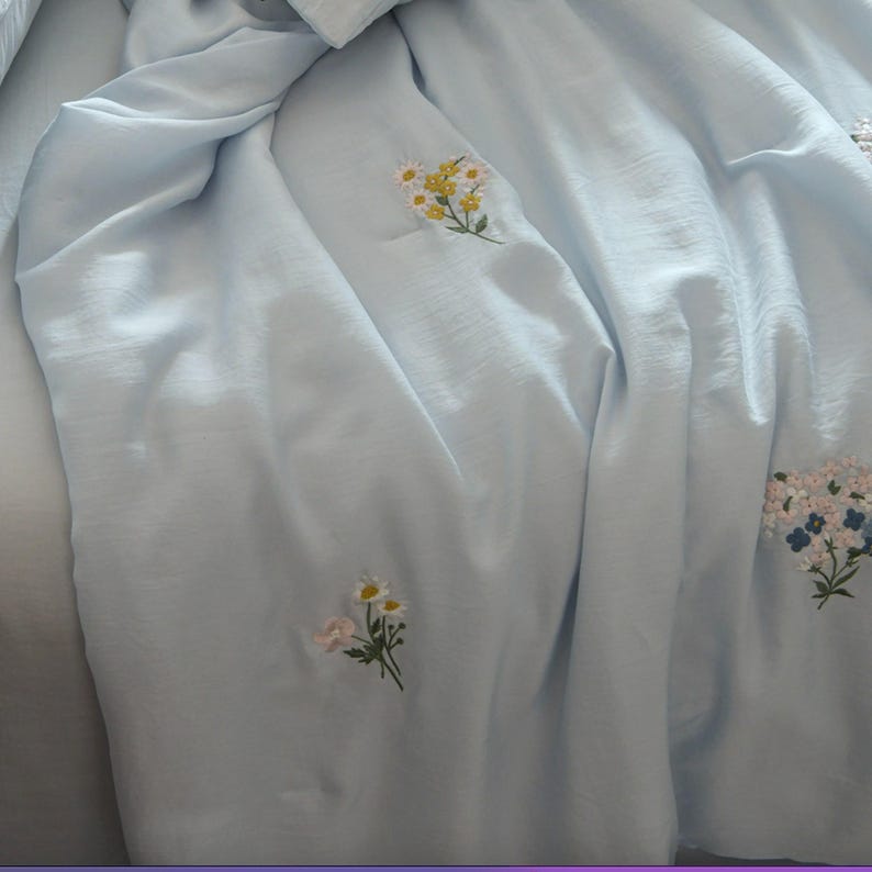 May include: A light blue fabric sheet or blanket featuring scattered embroidered floral motifs. The embroidery consists of small clusters of white, yellow, and pink flowers with green stems and leaves, distributed across the smooth, draped textile surface.