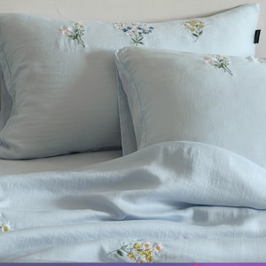May include: A light blue bedding set consisting of two rectangular pillowcases and a matching duvet cover. The fabric features delicate, embroidered floral clusters in shades of white, yellow, and blue scattered across the soft blue surface.