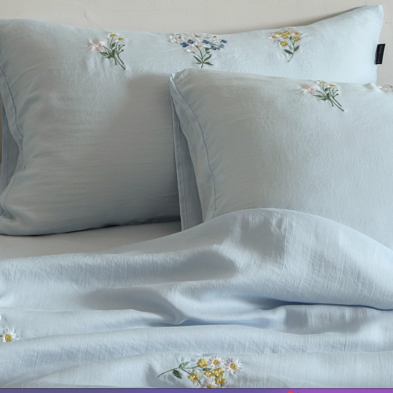 May include: A light blue bedding set consisting of two rectangular pillowcases and a matching duvet cover. The fabric features delicate, embroidered floral clusters in shades of white, yellow, and blue scattered across the soft blue surface.