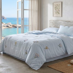 May include: A light blue duvet cover and matching pillowcase set featuring scattered, small, multi-colored floral embroidery patterns. The bedding is draped over a bed with a light-colored wooden headboard, positioned near a window overlooking the sea.